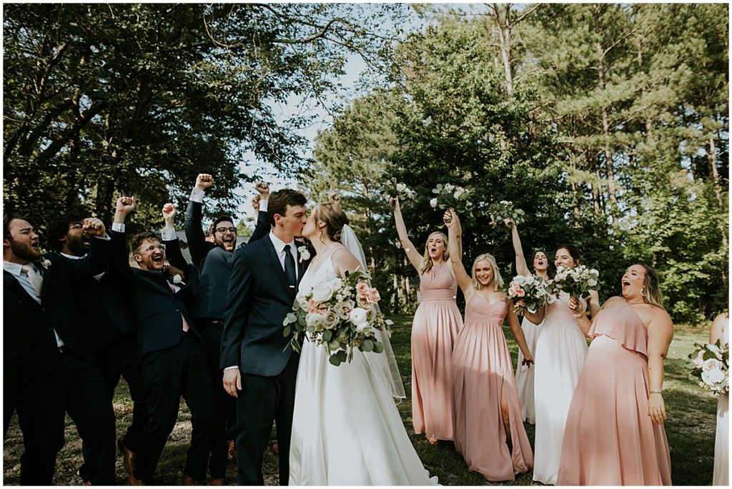 Bride and groom kiss with bridal party looking on and cheering! Summer wedding at rustic Auburn wedding venue