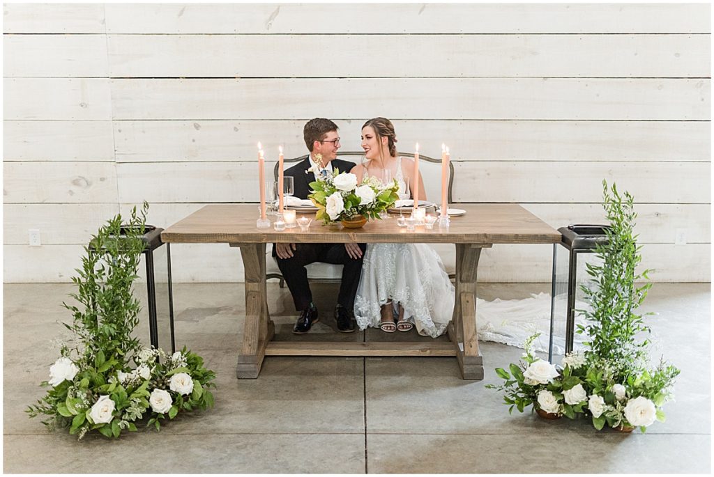 Bride and groom sat at a table with green and ivory florals and peach candles.
