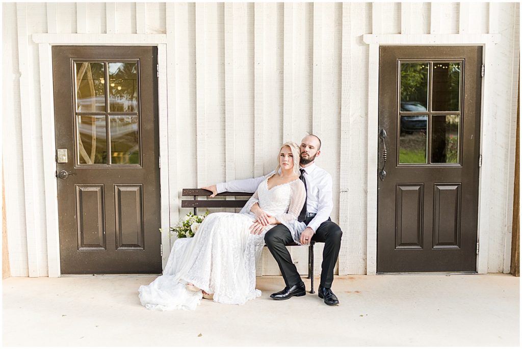 Bride and groom outside sitting on a bench with 2 brown doors either side at rustic elegant white barn wedding