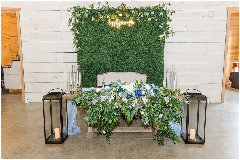 Sweethard table for bride and groom with greenery back drop, neon lights and blue and green florals and large candles.