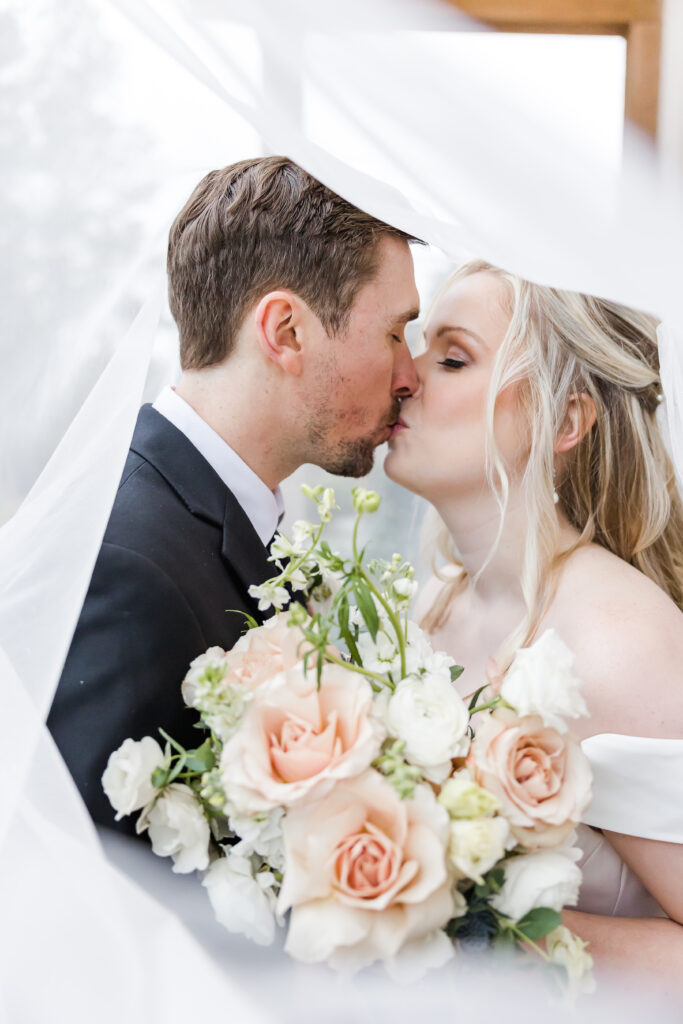Bride and groom under a veil holding florals and kissing.