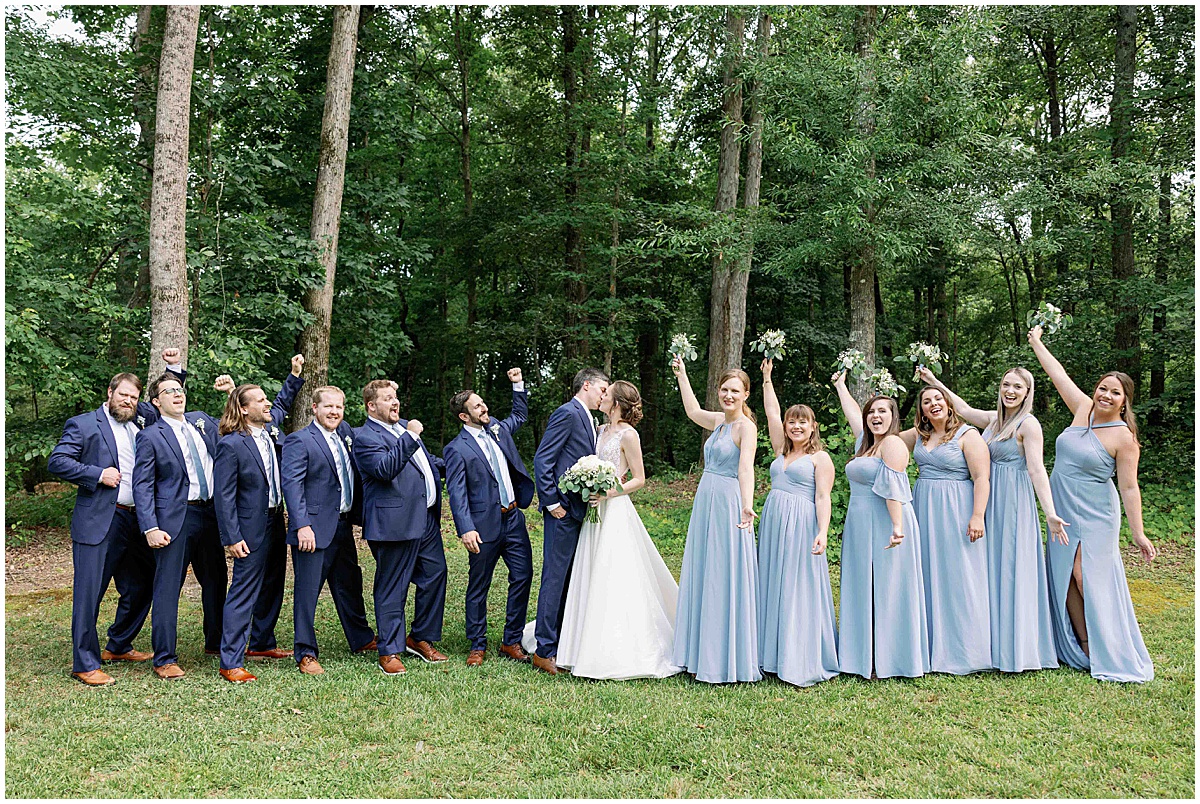 bridesmaids in light blue dresses and groomsmen in navy suits cheering bride and groom as they kiss.