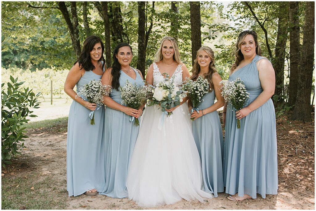 bride with bridesmaids wearing baby blues dresses at forest area at Koury Farms