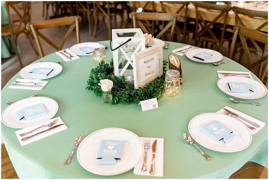 wedding table with green linen, lantern, candles, green wreath and drink coozy at each place setting